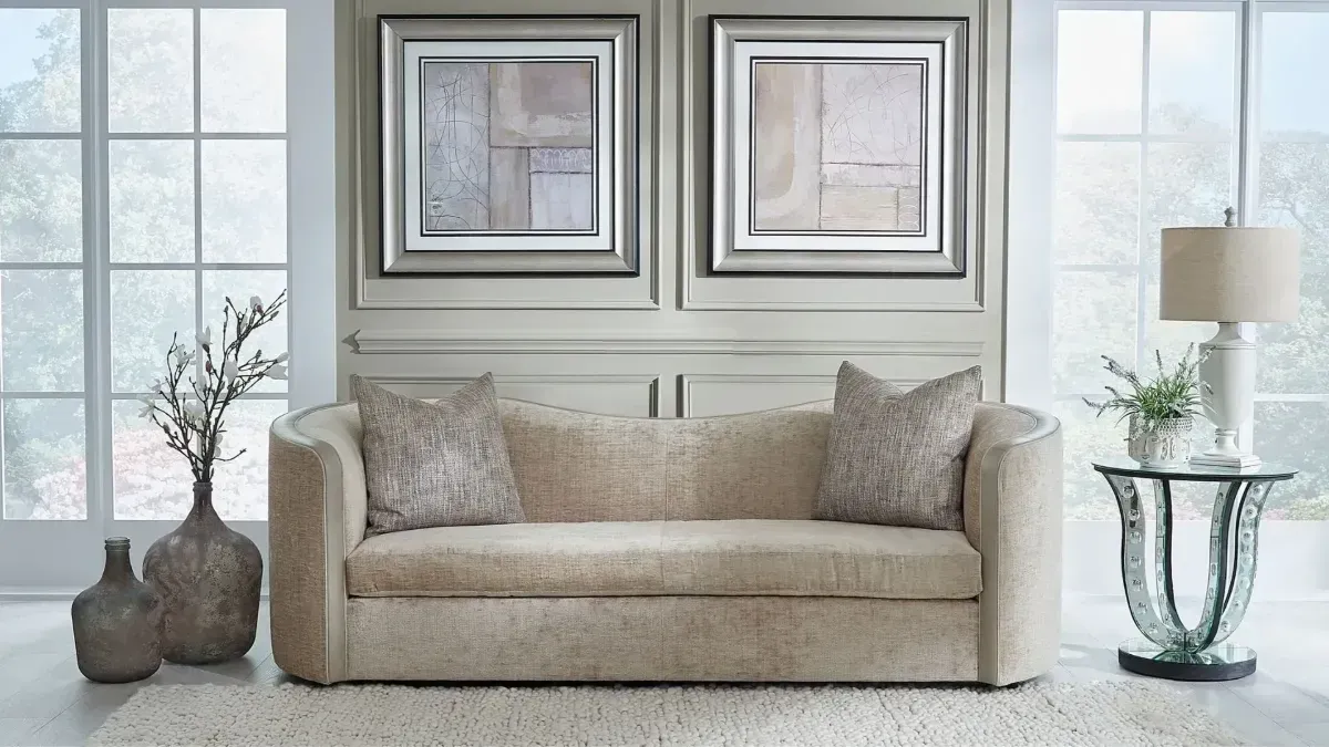 Bright living room with a beige sofa, framed wall art, lamps, and decorative vases by windows.
