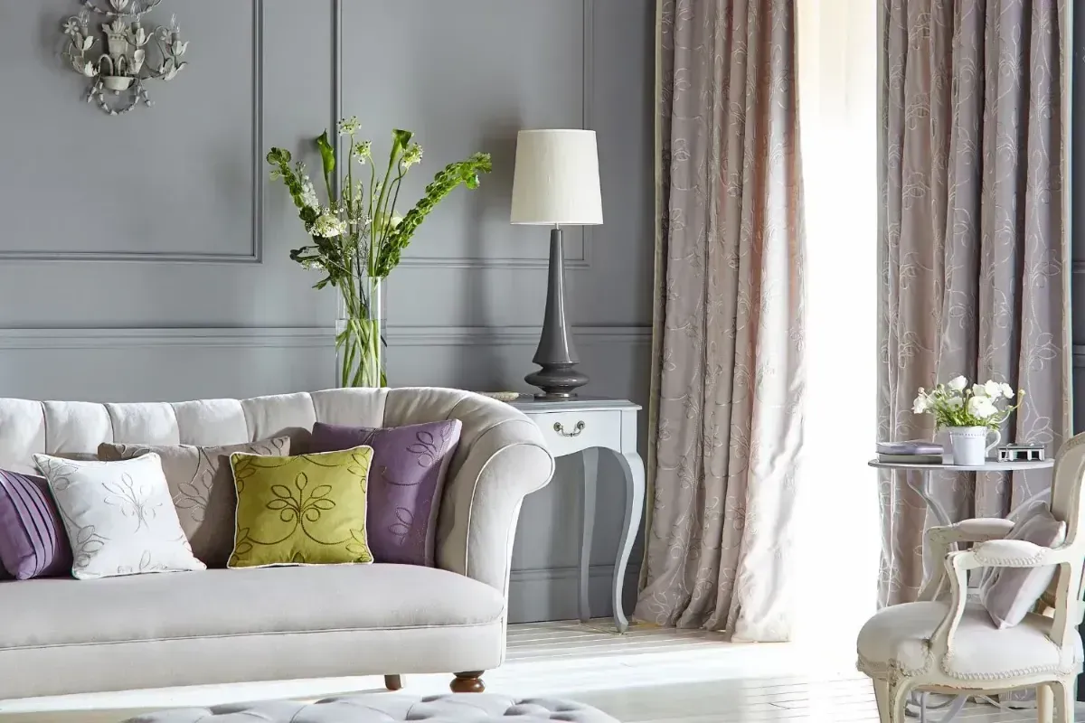 Elegant living room with white sofa, purple and green pillows, gray walls, and tall curtains by a bright window