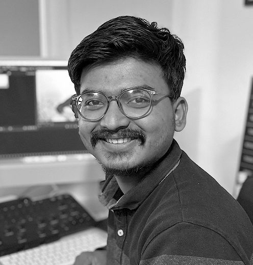 Smiling person with glasses seated at a desk in front of a computer monitor and keyboard