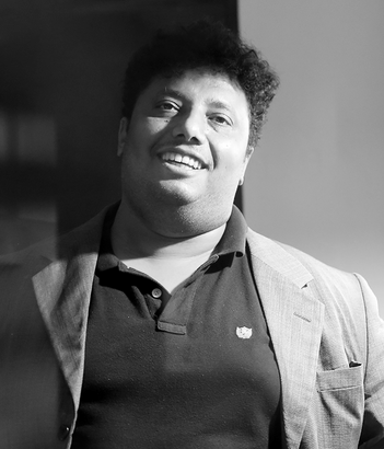 Person smiling in a blazer over a dark polo shirt, standing against a plain background.
