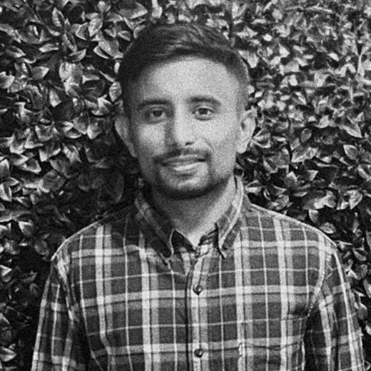 Smiling person in a plaid shirt standing before a leafy wall.