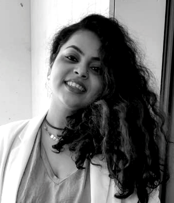 Smiling person with curly hair in a light jacket, posing indoors near a window.