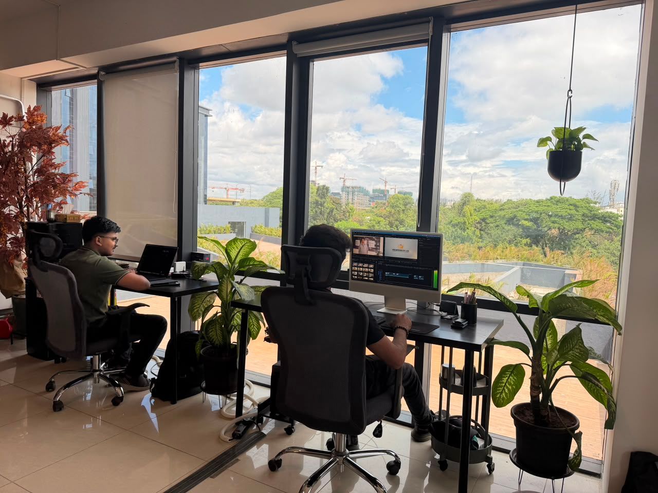 Two people working at desks by large windows in a bright office with city views and plants