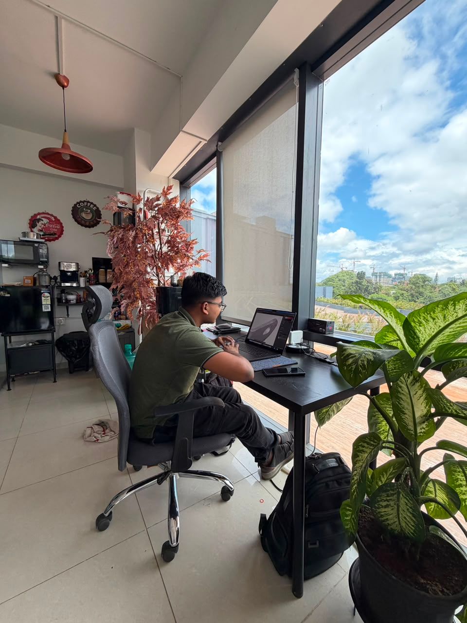 Person working on a laptop at a desk beside large windows in a bright office with plants and a city view