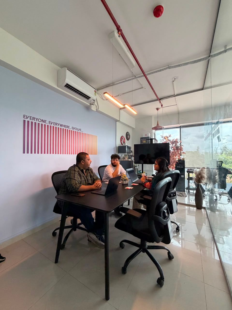 Three people meeting around a black conference table in a bright modern office, with a TV and glass wall.
