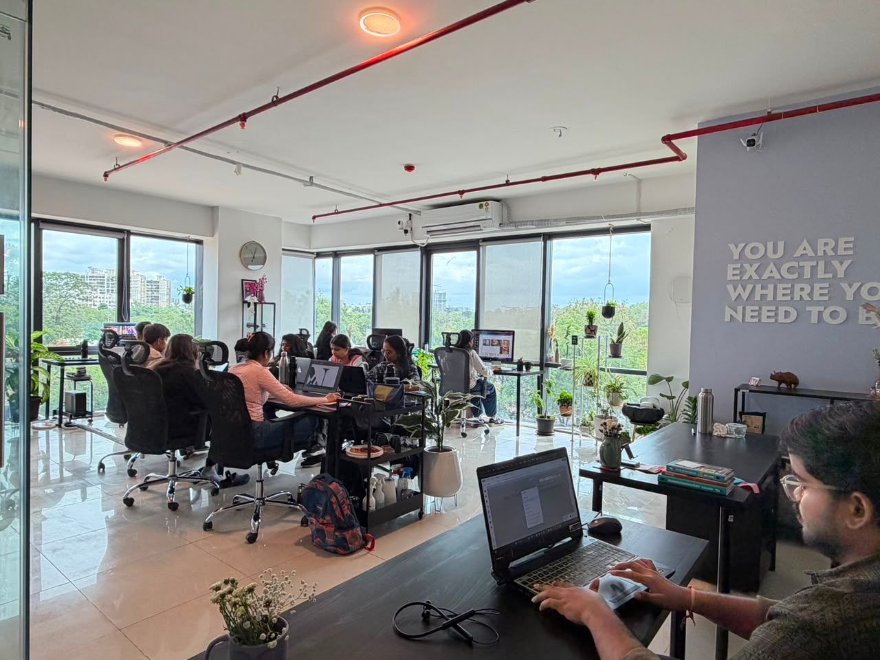 Open office with people working at desks near large windows and a wall sign saying “YOU ARE WHAT YOU NEED TO BE.”