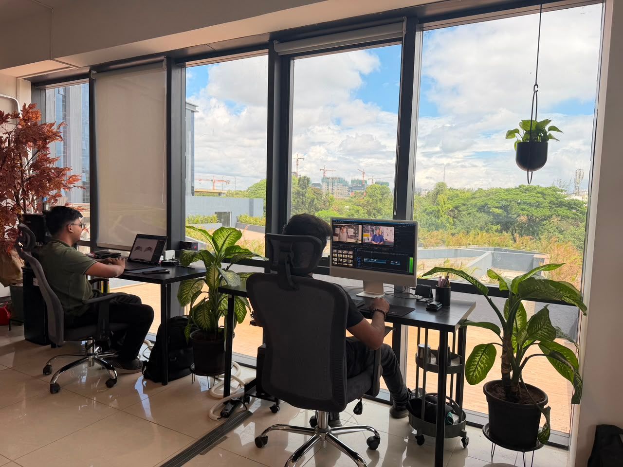 Two people work at desks in a bright office with large windows and many plants.