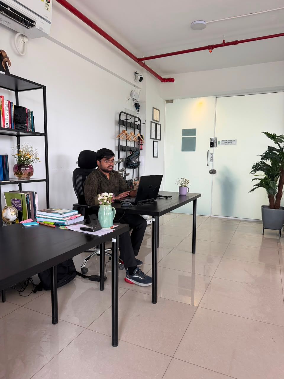 Office with two people at desks, laptop, shelves, potted plants, and white glass doors in the background