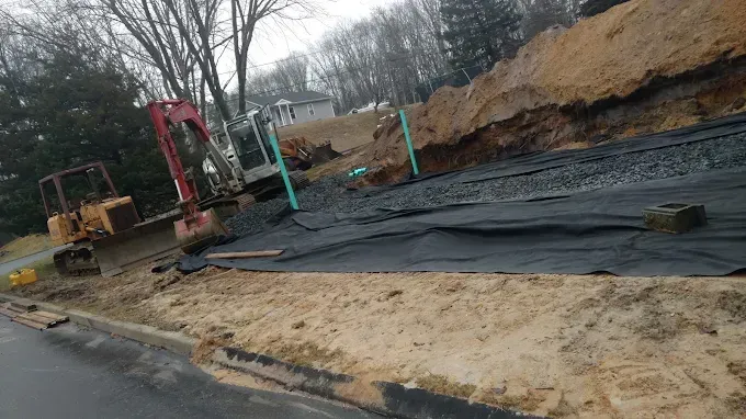 Construction site with excavator, black tarp, and dirt beside a wet roadside