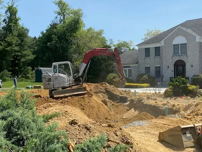 Excavator digging a dirt trench beside a large house on a sunny day