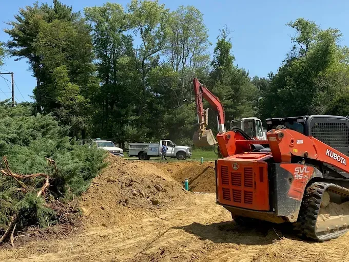 Orange excavator on a dirt construction site beside trees and a pickup truck under a blue sky.