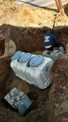 Worker crouches in a trench beside a large white utility tank under a dirt embankment.