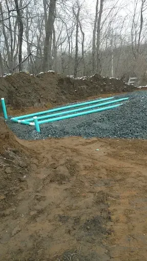 Blue PVC pipes laid on gravel beside a dirt trench at a wooded construction site