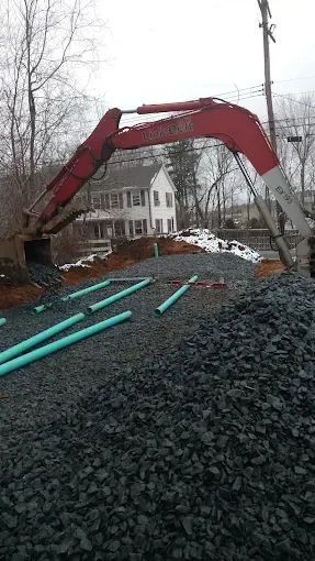 Red excavator on a dirt pile at a construction site, with houses and bare trees in the background.