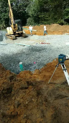 Construction site with excavator, gravel pile, and survey tripod on a gravel road.