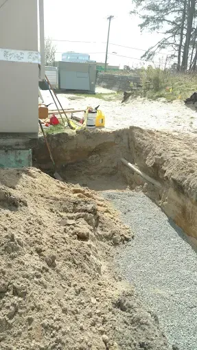 Open trench beside a building, filled with gravel and dirt at a construction site.
