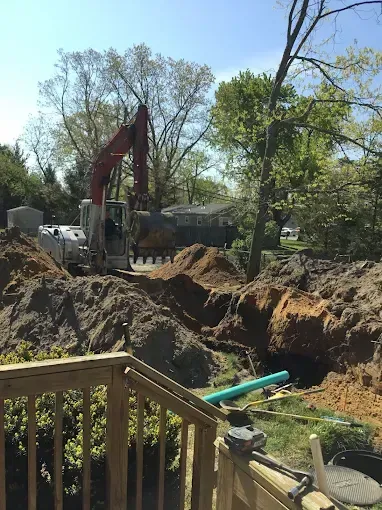 Excavator digging a large trench in a wooded backyard beside a house on a sunny day