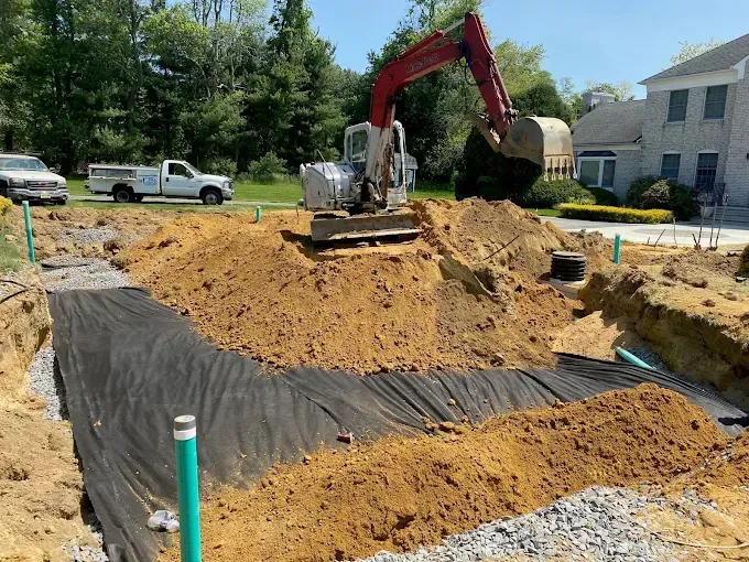 Excavator digging soil at a construction site beside a house and parked vehicles