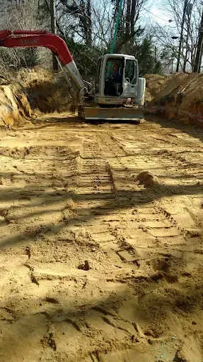 Excavator clearing a dirt path at a construction site, with a rough roadbed and trees in the background.