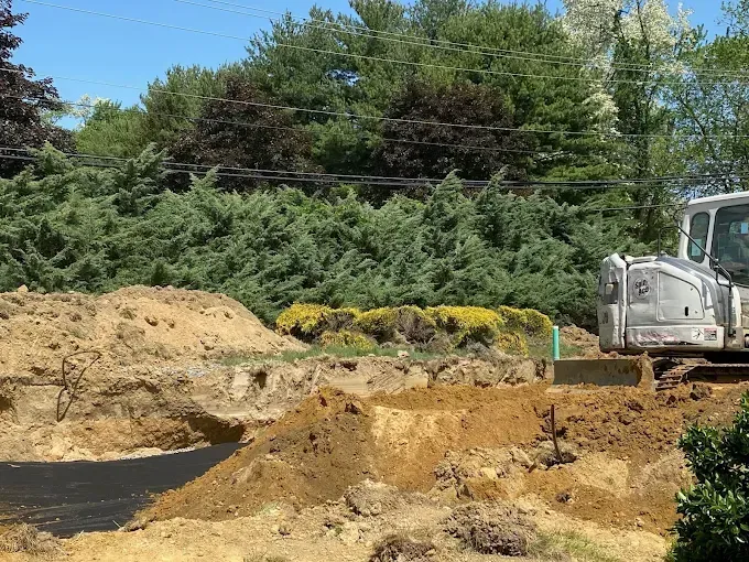 Construction site with dirt piles, excavator, and trees under a clear sky