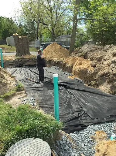 Worker standing in a trench lined with black plastic near dirt piles and trees.