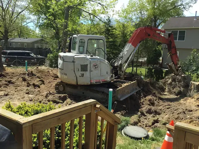 Excavator digging in a yard beside a deck and trees, with a house in the background.