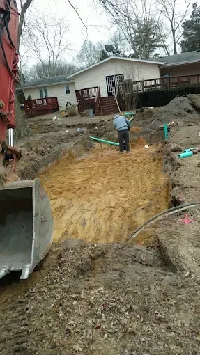 Backyard construction site with a deep dirt excavation and a worker standing near a house