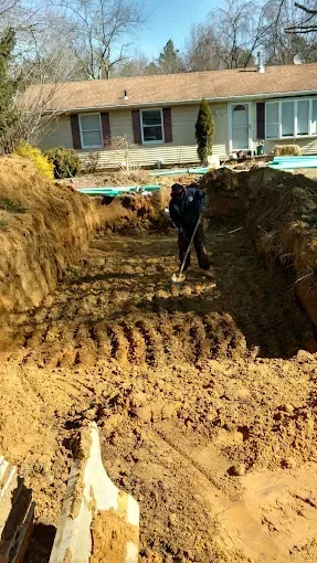 Worker digging a deep backyard excavation beside a house, with an excavator bucket in the foreground.