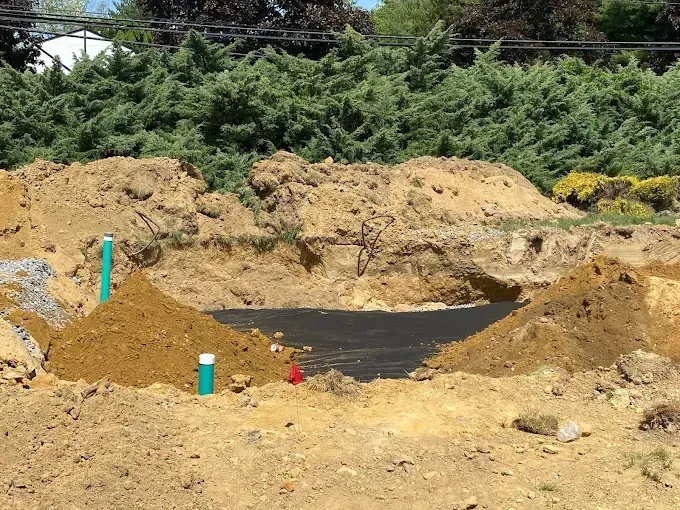 Excavation site with dirt mounds, black liner, and colored utility markers beside trees