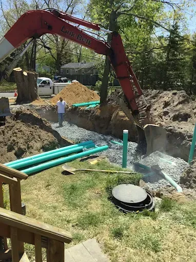 Excavator digging a trench beside green pipes and a worker in a yard.