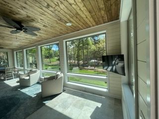 Modern sunroom with wood ceiling, wall-mounted TV, and beige chairs facing large windows and a green yard