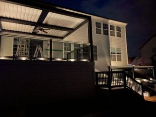 Night view of a lit porch deck and house exterior with railings and a fan under the roof.