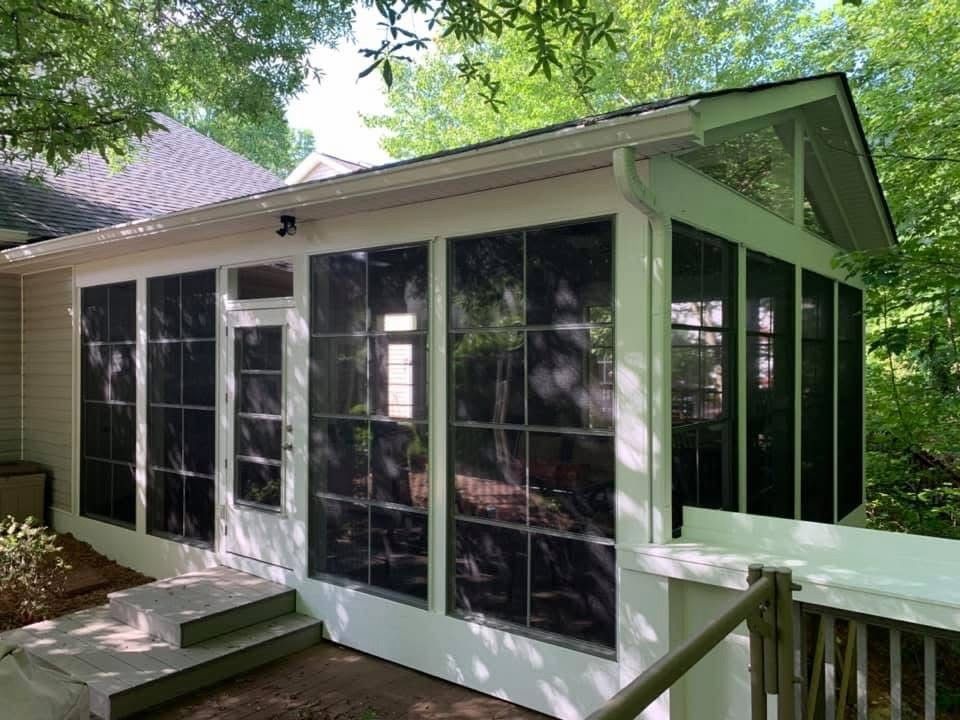 Screened porch with white-framed windows and doors beside a house, surrounded by trees.