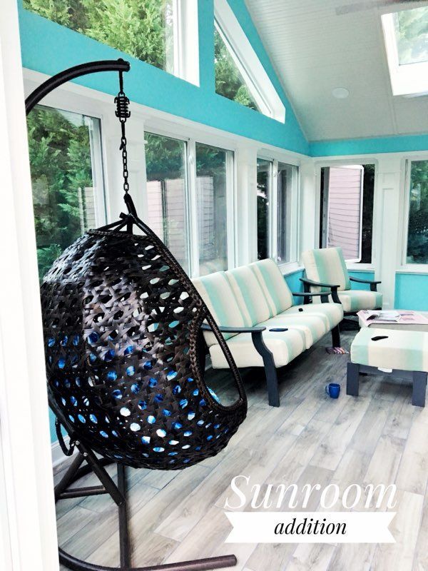 Bright sunroom with turquoise walls, white sofa, and black hanging chair by large windows.