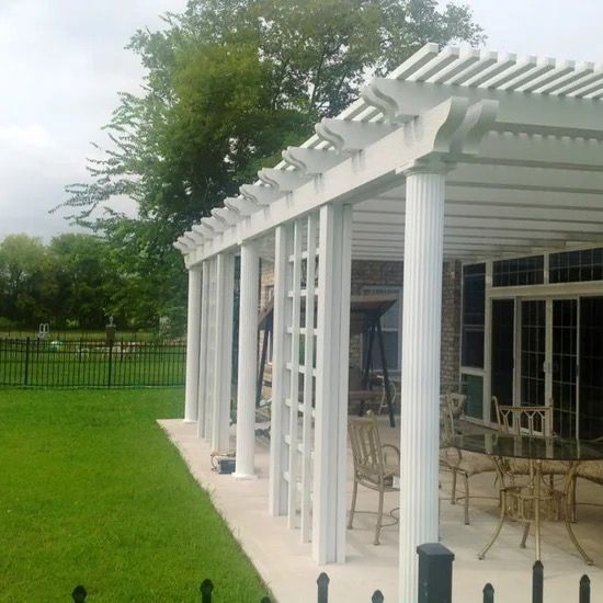 White pergola beside a patio with outdoor chairs, overlooking a green lawn and trees