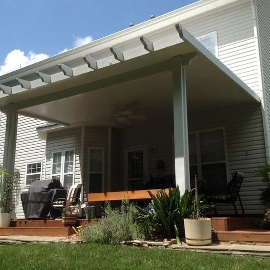 Covered patio with gray support posts, wooden deck, potted plants, and outdoor chairs on a house exterior