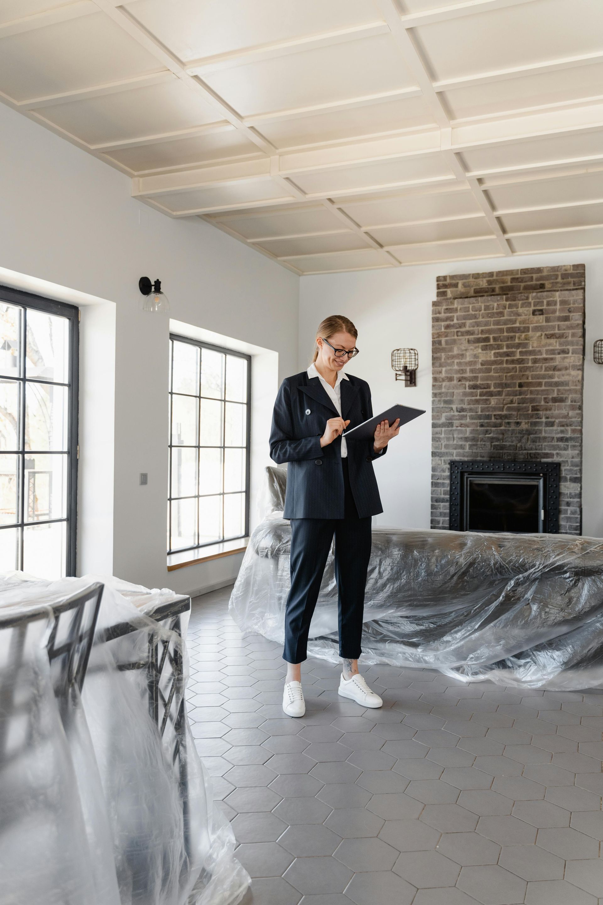 Woman in suit with tablet inspects a large room with covered furniture and a brick fireplace.