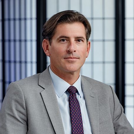 A professional portrait of a person in a gray blazer, light blue shirt, and patterned tie, set against a grid background.