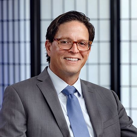 A person in a gray suit, light blue dress shirt, and blue tie, smiling against a blurred background with vertical lines.