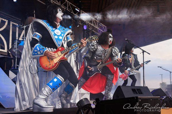 a group of people playing guitars on a stage with a kiss logo in the background