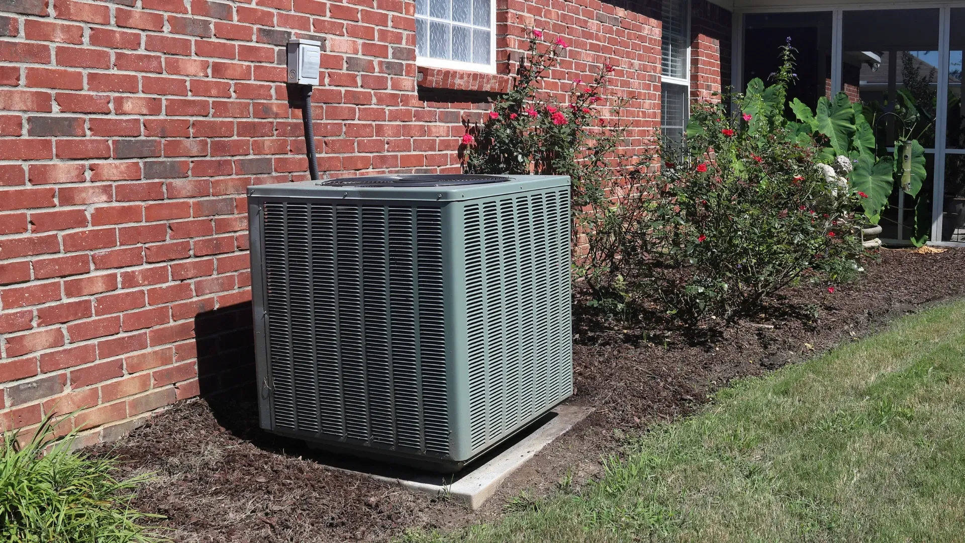 Air conditioning unit next to a brick wall and landscaping.