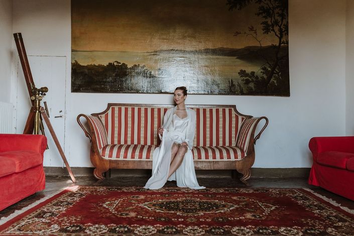 Una donna in abito bianco è seduta su un divano a righe rosse in un'elegante stanza con un dipinto paesaggistico.