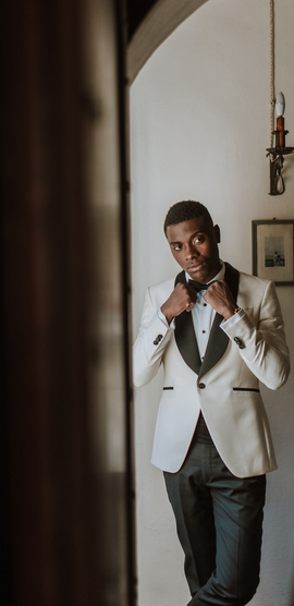 Man adjusting his tie in a white tuxedo jacket by a doorway, with a framed picture on the wall.