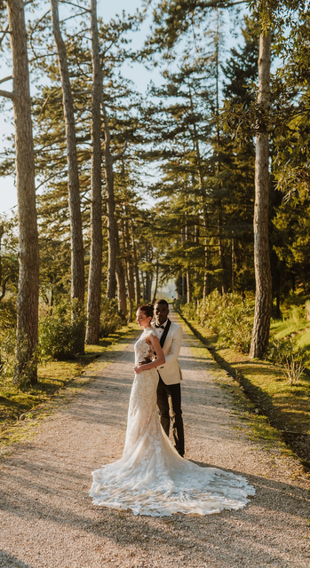 Bride and groom standing on a forest path, embracing under tall pine trees
