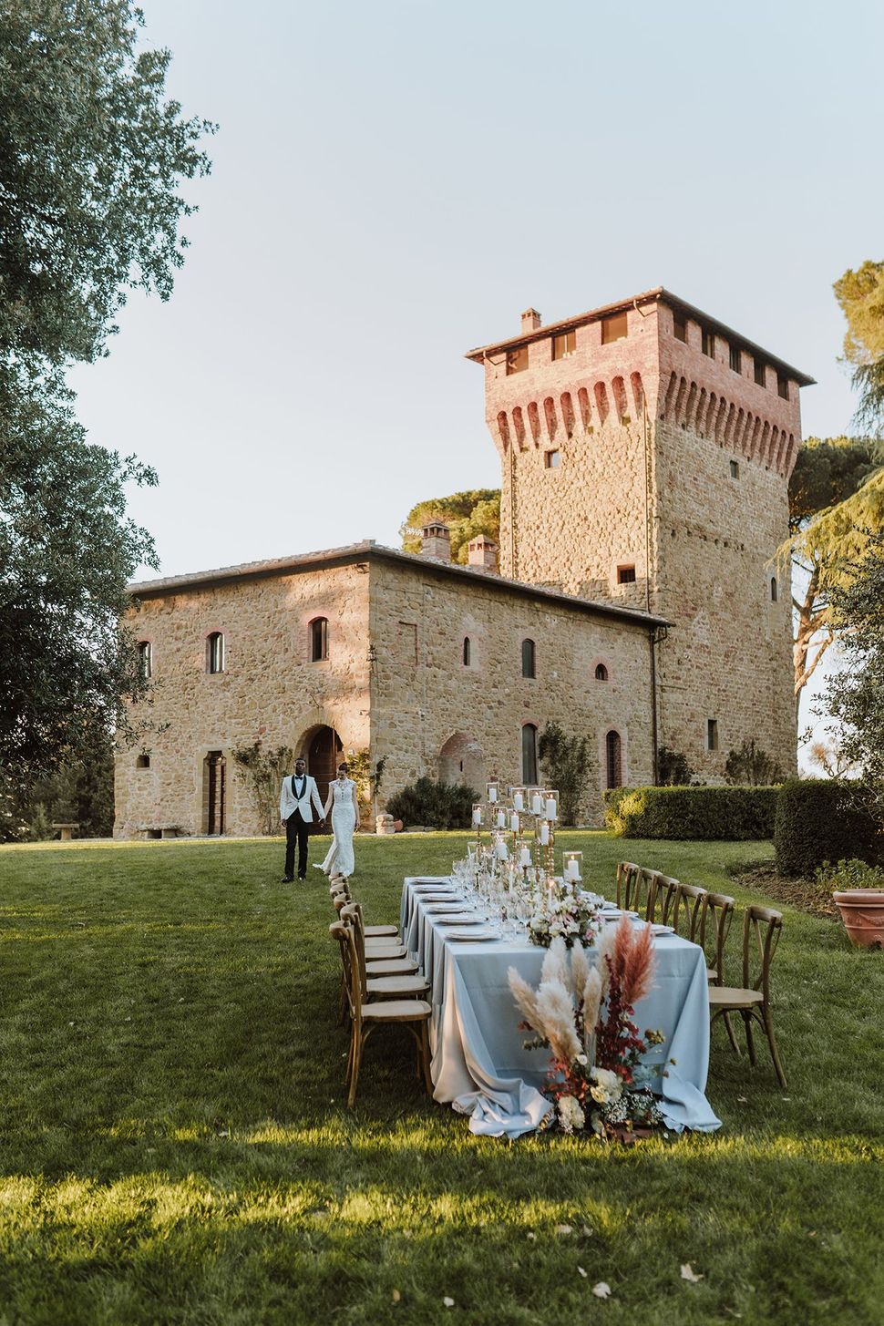 Tavolo per ricevimento di nozze all'aperto allestito su un prato accanto alla torre di pietra di un castello al tramonto