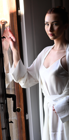 Una donna in abito bianco è in piedi accanto a una finestra e guarda fuori con un'espressione serena.