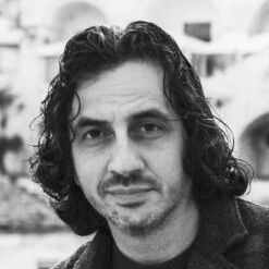 Black-and-white close-up portrait of a man with shoulder-length curly hair and a neutral expression