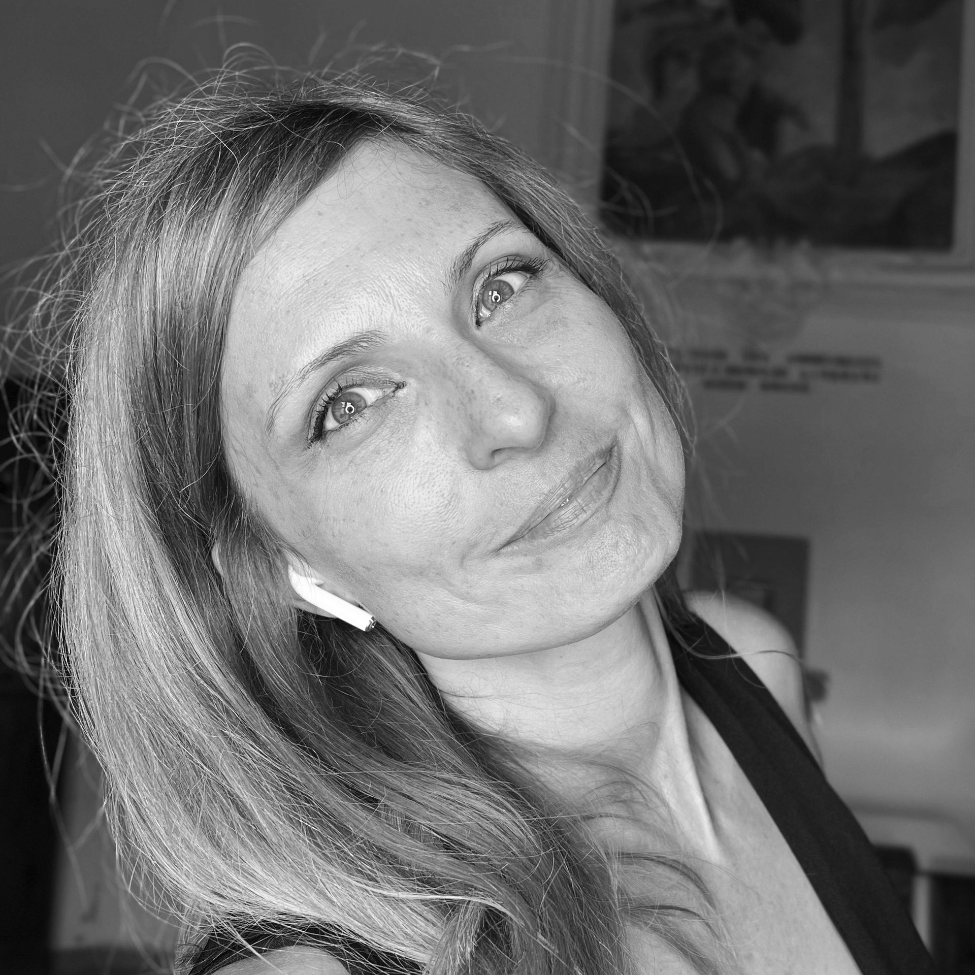 Black-and-white close-up of a smiling person with long hair, tilting their head indoors.