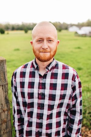 A man with a beard is wearing a plaid shirt and standing in a field.