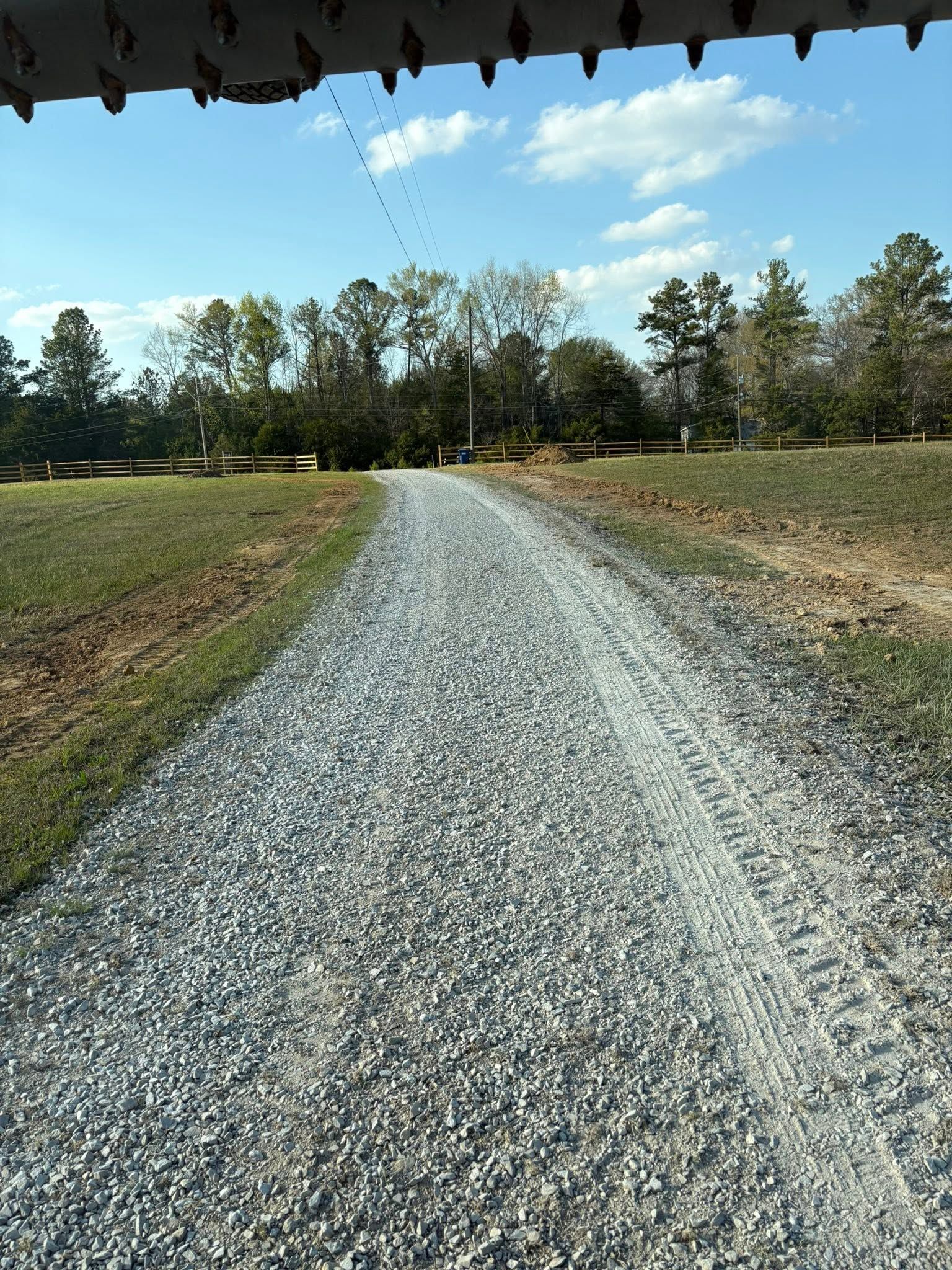 Gravel road stretching through grassy fields toward a tree line under a blue sky with clouds